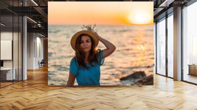 Young beautiful woman in dress and hat holding a bouquet of flowers at sunset by the sea, romantic mood Wall mural