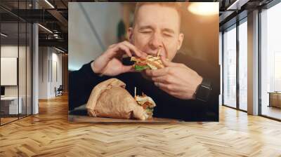 Handsome man sitting in a restaurant and eating club sandwich. He is taste that big delicious dish. Young guy having his lunch Wall mural