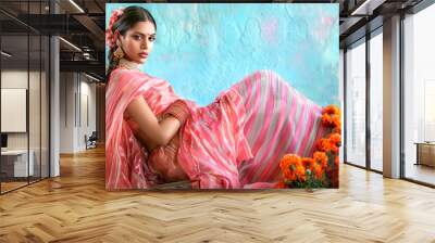 A fashion of an Indian model wearing a pastel pink linen sari with stripes, sitting on a chair in front of an ancient wooden cabinet and yellow marigold flowers Wall mural