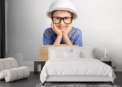 Smiling boy engineer in a construction helmet builds a house from wooden blocks on a table with plans and a measure tape. Child builder plays with cubes at home. Mortgage, dream, choice of profession. Wall mural