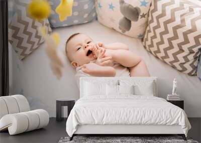 Cute baby lies in a white round bed. Light nursery for young children.  Toys for infant cot. Smiling child playing with mobile of felt and chewing his feet in sunny bedroom. Wall mural