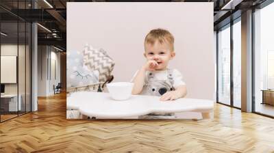 Cute baby sitting at the table and eating in the children's room bright colors. Wall mural