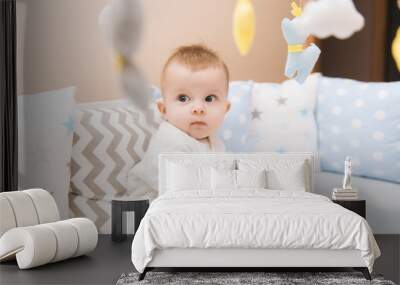 Cute baby sitting in a white round bed. Light nursery for young children.  Toys for infant cot. Smiling child playing with mobile of felt in sunny bedroom. Wall mural