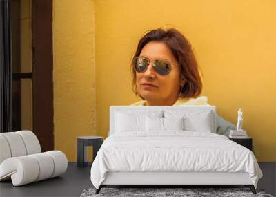 Confident woman in sunglasses relaxing against yellow wall in casual attire Wall mural