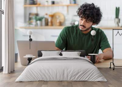 Concentrated young Asian man student using laptop computer for online education, taking notes in a notebook, studying, distance learning at home Wall mural