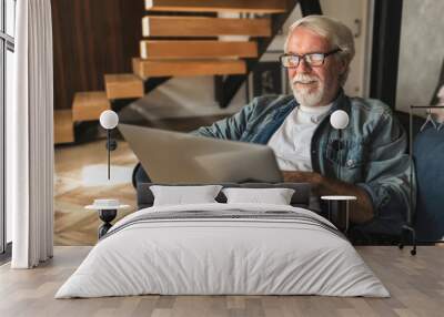 Elderly man with glasses with gray hair and beard sits at home on the sofa works or browses social networks on laptop. Happy carefree pension Wall mural