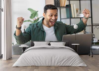 Excited young Indian man in casual clothes celebrating dream goal achievement. Successful businessmen rejoices financial project results sitting at the desk in the home office Wall mural