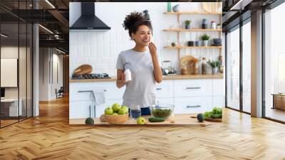 Happy african american woman standing at the cuisine table in the home kitchen drinking dietary supplements, looking away and smiling friendly, healthy lifestyle concept Wall mural