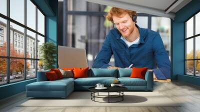 Happy male call center worker with headset consults a client. Redhead man freelancer or student using laptop for remote work from home. Learning, education remotely online, studying Wall mural