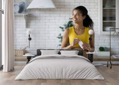 Happy sportive african american woman in sportswear holding a bottle of nutritional supplements, looking happily out the window, healthy lifestyle Wall mural