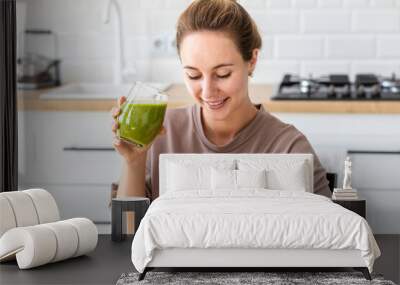 Healthy lifestyle, diet eating, weight loss concept. Young woman drinking fresh green smoothie communicates or browsing in social media using a mobile phone sitting at home in the kitchen Wall mural