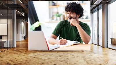 Indian young man using laptop for watching webinar, studying online and takes notes in a notebook, male student is studying online, e-learning concept Wall mural