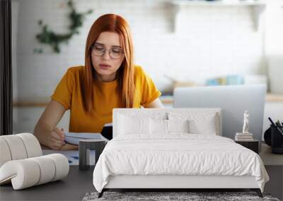 Online learning, portrait of focused beautiful red-haired young female student with laptop and notepad sitting at the table, distance education Wall mural