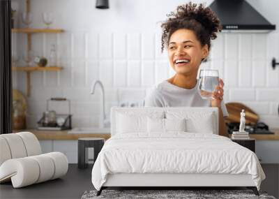 Portrait of a beautiful African American young woman drinking water standing in the kitchen at home looking away and smiling, wellness healthy food concept Wall mural