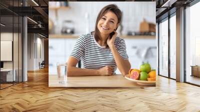 Portrait of a beautiful woman with a glass of water and fruits sitting at a table in the kitchen looking at the camera and smiling , wellness healthy food concept Wall mural