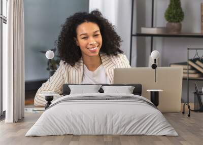 Portrait of a young successful African American businesswoman or an office worker sitting at a desk with a laptop in a modern office, looking at the camera and smiling Wall mural