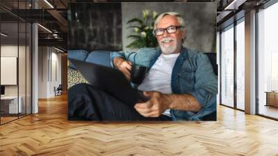 Portrait of smiling kind elderly man sitting on the couch with cup of coffee and laptop computer. Happy pension Wall mural