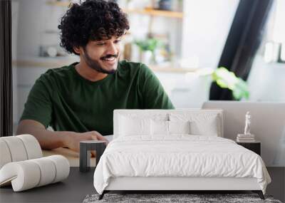 Smiling Indian young man using laptop for watching webinar, studying online and takes notes in a notebook, male student is studying online, e-learning concept Wall mural