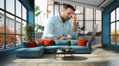 Tired Indian young man feel eye strain from working at the computer for a long time sitting at a desk in a home office Wall mural