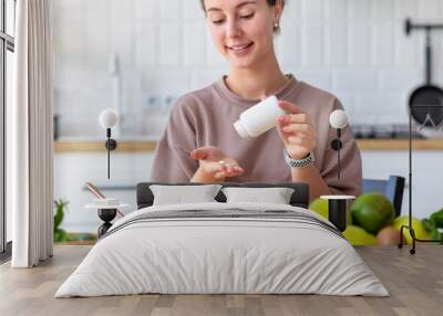 Vitamins, nutritional supplements, healthy lifestyle concept. Young woman eating breakfast and taking vitamin sitting at home in the kitchen, smiling friendly Wall mural