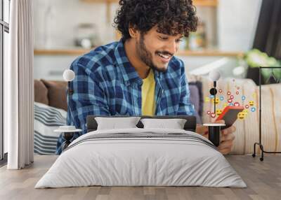 Young happy Asian man using social media with a mobile phone. Smiling male student with curly hair looking through likes, sitting on the couch at home Wall mural