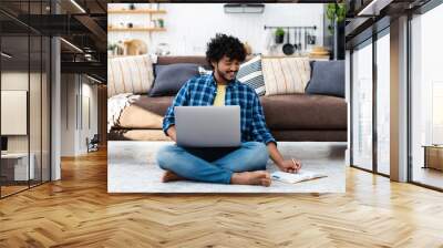 Young Indian male student studying remotely at home, online learning. Successful man freelancer sitting on the floor uses laptop for work on the Internet Wall mural