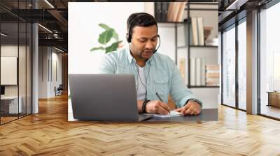 Young indian man in headset taking notes in notebook while watching online webinar using laptop computer, online studying at home concept education Wall mural