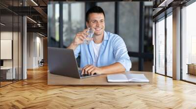 Young successful businessman man drinking clean water sitting at the office table. Smiling freelancer using laptop for working,  holding glass of fresh water in his hand Wall mural