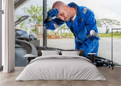 A tired male car mechanic wiping off his sweat from his forehead while working on car's engine at his car repair garage Wall mural