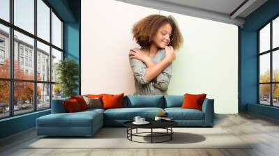 african american little girl feeling in love, smiling, cuddling and hugging self, staying single, being selfish and egocentric against flat wall Wall mural