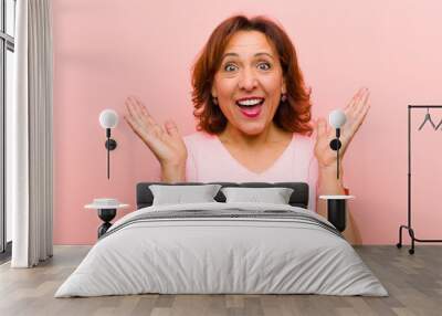 middle age woman looking happy and excited, shocked with an unexpected surprise with both hands open next to face against pink wall Wall mural