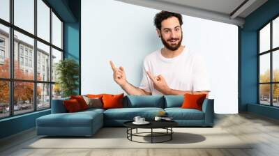 young adult bearded man smiling happily and pointing to side and upwards with both hands showing object in copy space Wall mural