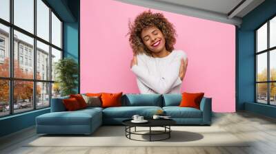 young african american woman feeling in love, smiling, cuddling and hugging self, staying single, being selfish and egocentric against pink wall Wall mural