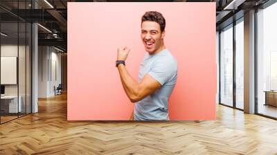 young arabian man feeling happy, satisfied and powerful, flexing fit and muscular biceps, looking strong after the gym against pink wall Wall mural