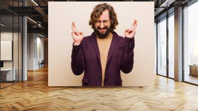 young bearded crazy man feeling nervous and hopeful, crossing fingers, praying and hoping for good luck against flat color wall Wall mural