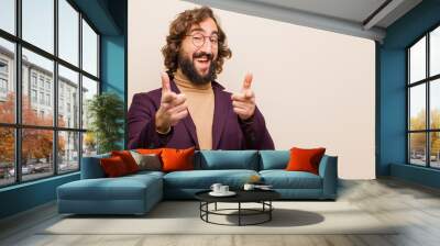 young bearded crazy man smiling with a positive, successful, happy attitude pointing to the camera, making gun sign with hands against flat color wall Wall mural