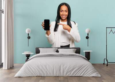 young black woman smiling cheerfully, feeling happy and pointing to the side and upwards, showing object in copy space. smart phone concept Wall mural