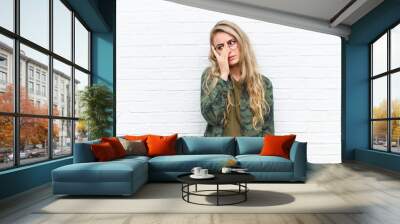 young blonde woman feeling bored, frustrated and sleepy after a tiresome, dull and tedious task, holding face with hand against brick wall background Wall mural