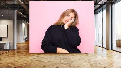 young blonde woman feeling bored, frustrated and sleepy after a tiresome, dull and tedious task, holding face with hand against flat wall Wall mural