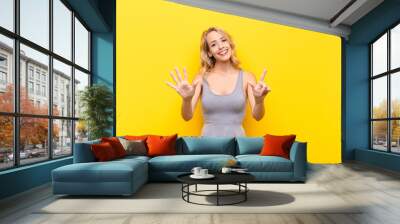 young blonde woman smiling and looking friendly, showing number seven or seventh with hand forward, counting down against orange wall Wall mural