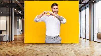 young handosme man smiling and feeling happy, cute, romantic and in love, making heart shape with both hands against flat color wall Wall mural