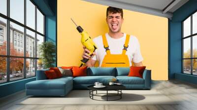 young handsome man with cheerful and rebellious attitude, joking and sticking tongue out. handyman and drill concept Wall mural