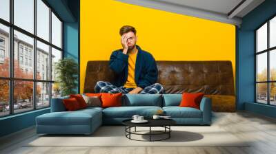 young man wearing pajamas feeling bored, frustrated and sleepy after a tiresome, dull and tedious task, holding face with hand . sitting on a sofa Wall mural