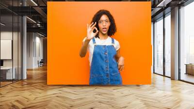 young pretty black woman feeling successful and satisfied, smiling with mouth wide open, making okay sign with hand against orange wall Wall mural