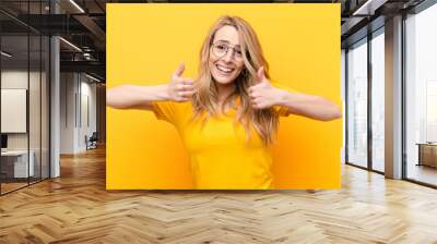 young pretty blonde woman smiling broadly looking happy, positive, confident and successful, with both thumbs up against flat color wall Wall mural