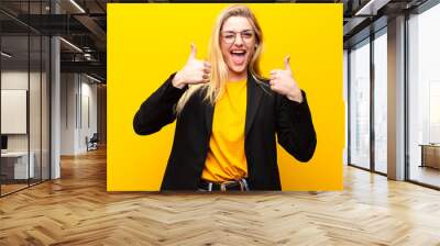 young pretty blonde woman smiling broadly looking happy, positive, confident and successful, with both thumbs up against yellow wall Wall mural