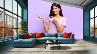 young pretty hispanic woman smiling cheerfully and pointing to copy space on palm on the side, showing or advertising an object Wall mural