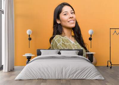 young pretty hispanic woman smiling to camera with crossed arms and a happy, confident, satisfied expression, lateral view against brown wall Wall mural