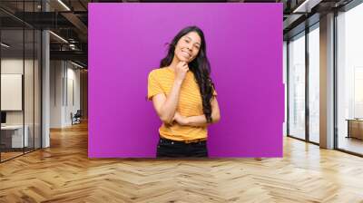 young pretty latin woman smiling, enjoying life, feeling happy, friendly, satisfied and carefree with hand on chin against purple wall Wall mural