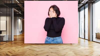 young pretty woman feeling scared or embarrassed, peeking or spying with eyes half-covered with hands against pink wall Wall mural